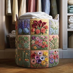 May include: A round, quilted laundry hamper with a floral patchwork design. The hamper features embroidered panels with various flowers and succulents in vibrant colors. It has two handles and is filled with a folded white blanket.