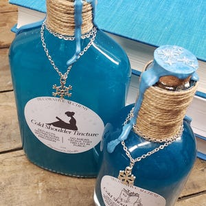 May include: Two blue glass bottles with a silver snowflake pendant necklace. The bottles are labeled "Cold Shoulder Tincture" and have a blue wax seal. The bottles are decorated with twine and are on a wooden surface.