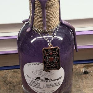 May include: A decorative purple glass bottle with a rope-wrapped neck and a silver chain. The bottle is sealed with purple wax and has a pendant with a sun and moon design. The label reads "Dreamless Sleep Draught".