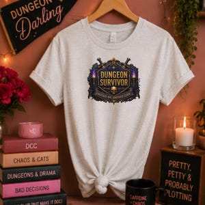 May include: A heathered light grey short-sleeve t-shirt featuring a central graphic with the text "DUNGEON SURVIVOR" and "ANOTHER DAY, ANOTHER FLOOR" above a skull, flanked by two swords and two lit torches on a dark blue shield-like emblem.