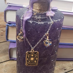 May include: A purple, textured bottle with a decorative chain and charms. The bottle's neck is wrapped in twine and sealed with purple wax. The chain holds a tarot card pendant and two crystal charms. The tarot card reads "The Wheel of Fortune."