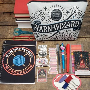 以下が含まれることがあります： 「YARN-WIZARD」の文字が入ったジッパー付きポーチ、 「If I'm not reading I'm crocheting」と書かれたノート、ステッカー、ペン、赤いメモ帳など、クラフトと読書用品のコレクション。 青いハサミと赤いペーパークリップも表示されています。