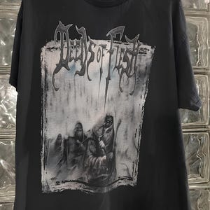 May include: Black t-shirt with the band name "Deeds of Flesh" in a gothic font above a grayscale graphic. The graphic shows hooded figures in a desolate setting. The shirt is made of a soft material.