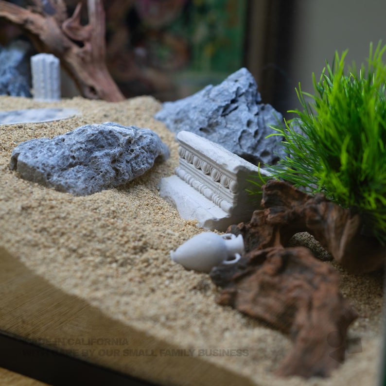 Pu&ograve; includere: Un paesaggio in miniatura per acquario con sabbia beige, rocce grigie strutturate, un frammento architettonico bianco con dettagli intagliati, una piccola anfora bianca, legno scuro e una pianta in plastica verde brillante. Testo: MADE IN CALIFORNIA.