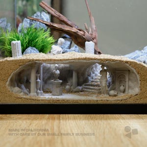 May include: A miniature diorama featuring a subterranean grotto with sandy textures, classical white columns, a bust on a pedestal, a standing figure, and small amphorae. The scene includes a stone staircase and is topped with green foliage and driftwood.
