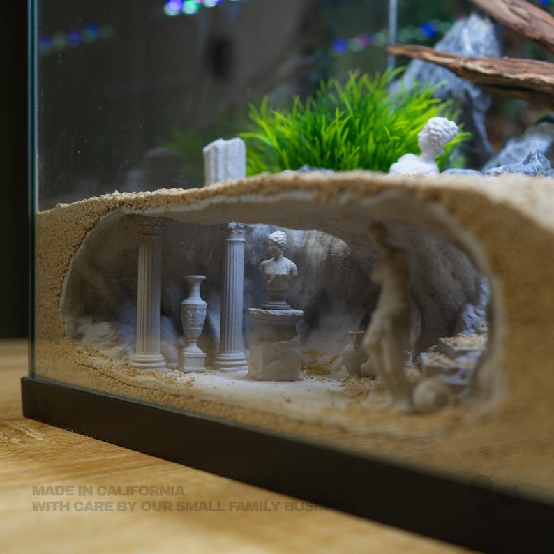 Pu&ograve; includere: Un acquario in vetro con un interno a grotta coperto di sabbia, caratterizzato da rovine bianche in miniatura in stile greco-romano, tra cui due colonne, un vaso decorativo, un busto centrale su piedistallo e una figura, con piante verdi.