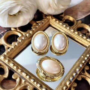 May include: A set of vintage gold-toned jewellery, including a pair of oval pearl earrings and a matching ring. The earrings and ring feature a large, lustrous white pearl set within a decorative gold frame. The jewellery is displayed on a mirrored surface.
