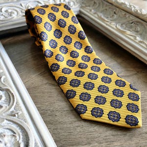 May include: A yellow tie featuring a repeating pattern of blue and gold floral medallions. The tie has a striped texture and is displayed on a wooden surface. A classic accessory for formal or business wear.