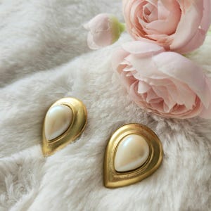 May include: Two gold-coloured earrings, each featuring a pearlescent, teardrop-shaped centre. The earrings are displayed on a soft, white, textured surface, with pink roses in the background. Jewellery.