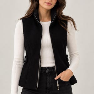 May include: A black sleeveless waistcoat featuring a high collar, a full-length silver-toned front zip, and two decorative side tabs with buttons. The waistcoat is worn over a white long-sleeved ribbed top and paired with dark charcoal denim jeans.