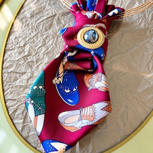Peut inclure: Une cravate en soie bordeaux ornée d'un motif répétitif de chaussures habillées colorées en bleu, vert et orange. L'accessoire est froncé sur le haut et maintenu par une broche circulaire dotée d'une pierre centrale sombre et d'un bord perlé.