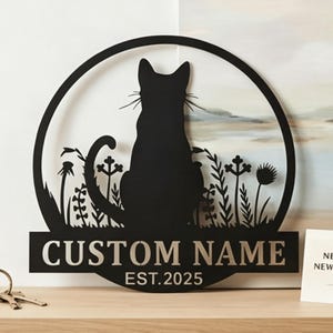 May include: Black metal cat silhouette wall art with a circular design. The cat is seated, facing away, within a circle with floral accents. The text "CUSTOM NAME" and "EST. 2025" are displayed on a lower rectangular bar.