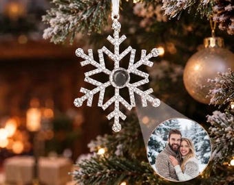 May include: A silver-toned snowflake ornament encrusted with clear crystals, featuring a central circular projection lens that displays a photograph of a couple in a snowy outdoor setting, suspended by a white ribbon from a pine branch.