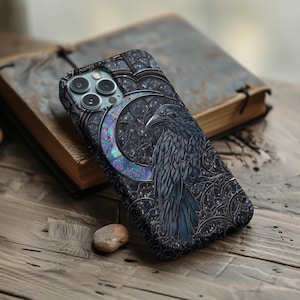 以下が含まれることがあります： 三日月を背景にカラスが描かれたスマホケース。ステンドグラス風のデザインで、濃い青、黒、虹色のアクセントが特徴です。木製の表面に置かれ、古びた本の隣に配置されています。