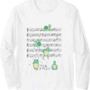 May include: A white long-sleeve t-shirt featuring a graphic print of five light green turtles interacting with black musical notes on a staff. The turtles are depicted in various poses, with some appearing to play or dance among the musical notation.