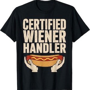 May include: A black short-sleeved t-shirt featuring the text "CERTIFIED WIENER HANDLER" in a distressed beige font. Below the text is a graphic illustration of two hands holding a hot dog in a bun with a yellow line of mustard on top.