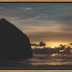 May include: A framed landscape cross-stitch depicting a coastal scene at sunset. A large, dark silhouette of a rock formation dominates the left side, with the ocean and a colorful sky filled with clouds and warm hues of orange and yellow.