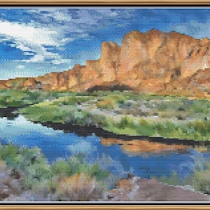May include: A framed cross-stitch landscape depicts a river reflecting a blue sky with fluffy white clouds. The river flows through a valley with green vegetation and is bordered by reddish-brown rock formations. The artwork is framed in a gold-colored frame.