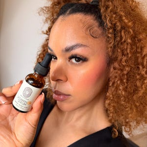 Puede incluir: Una botella de vidrio marr&oacute;n de suero de vitamina C es sostenida. La botella tiene un cuentagotas negro y una etiqueta blanca con texto y un logotipo. La persona tiene cabello casta&ntilde;o rizado y lleva una blusa negra.