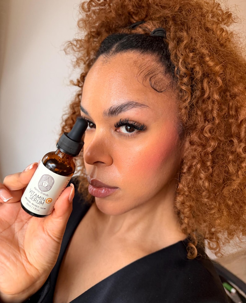 Puede incluir: Una botella de vidrio marr&oacute;n de suero de vitamina C es sostenida. La botella tiene un cuentagotas negro y una etiqueta blanca con texto y un logotipo. La persona tiene cabello casta&ntilde;o rizado y lleva una blusa negra.
