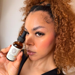 Op de afbeelding: Een bruine glazen fles met vitamine C-serum wordt vastgehouden. De fles heeft een wit etiket met zwarte tekst en een logo. De serumfles wordt voor het gezicht van een persoon met krullend bruin haar en roze blush gehouden.