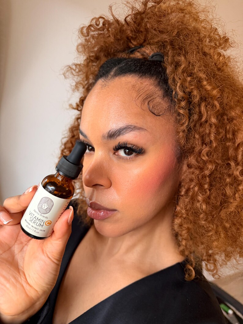 Op de afbeelding: Een bruine glazen fles met vitamine C-serum wordt vastgehouden. De fles heeft een wit etiket met zwarte tekst en een logo. De serumfles wordt voor het gezicht van een persoon met krullend bruin haar en roze blush gehouden.