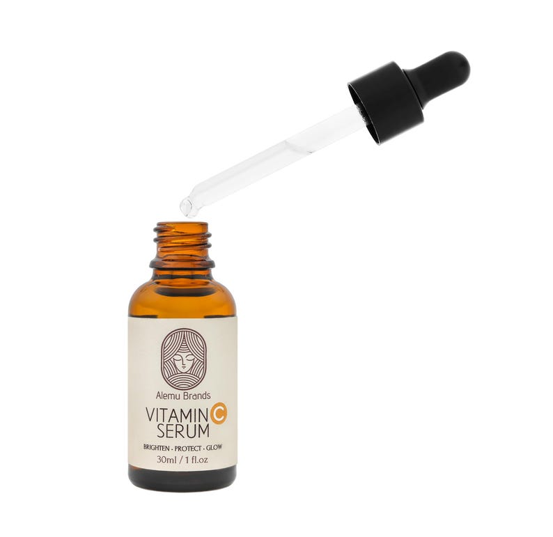 Op de afbeelding: Een bruine glazen fles vitamine C serum met een zwarte druppelaar. Het etiket luidt "Alemu Brands Vitamin C Serum" met de slogan "Brighten - Protect - Glow". De fles bevat 30 ml / 1 fl.oz.