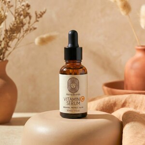 Op de afbeelding: Een bruine glazen fles vitamine C serum met een zwarte druppelaar. Het etiket bevat de merknaam "Alemu Brands" en de woorden "Vitamin C Serum". De fles is 30 ml / 1 fl.oz. en staat op een beige voetstuk.