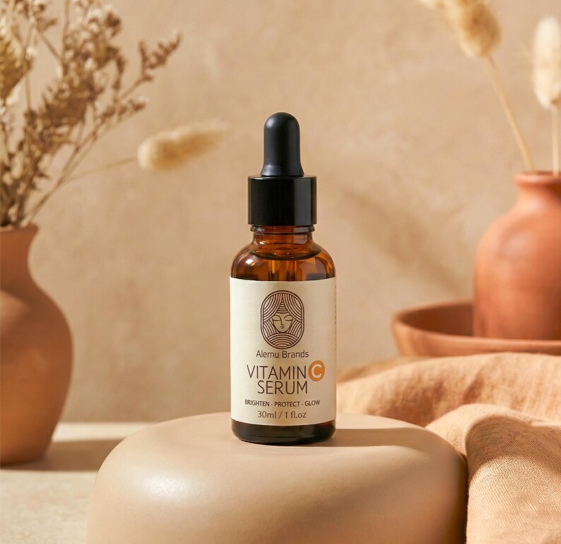 Op de afbeelding: Een bruine glazen fles vitamine C serum met een zwarte druppelaar. Het etiket bevat de merknaam "Alemu Brands" en de woorden "Vitamin C Serum". De fles is 30 ml / 1 fl.oz. en staat op een beige voetstuk.