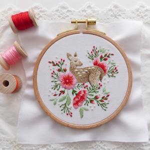 May include: A white embroidery hoop with a brown wooden frame. The hoop features a hand-stitched design of a fawn surrounded by pink and red flowers. The hoop is sitting on a white fabric with a lace trim.