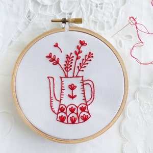 May include: Red embroidery of a teapot with flowers in a hoop. The teapot has a red floral pattern and the flowers are stylized.