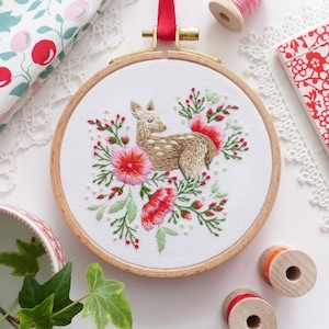 May include: Embroidered hoop art featuring a fawn surrounded by pink and red flowers. The hoop is made of wood and has a white fabric background.