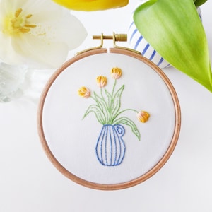 May include: Embroidered hoop art featuring a blue and white striped vase with a bouquet of yellow and orange tulips.