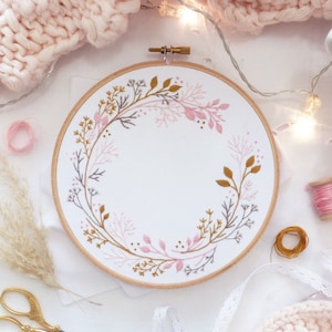 May include: A white embroidery hoop with a pink and gold floral design. The hoop is on a white surface with a pink knitted blanket and a strand of white lights.