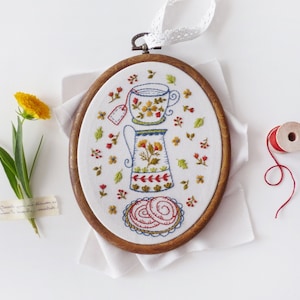 May include: Embroidered oval hoop art depicting a tea set with floral details. The design shows a teacup, teapot, and pastries, all decorated with flowers and leaves. The wooden frame is suspended by a white lace ribbon.
