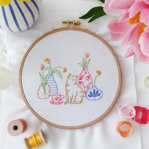 May include: Embroidered hoop art featuring a cat sitting in front of four colorful vases with flowers. The vases are decorated with floral patterns and geometric designs. The cat is orange and white.