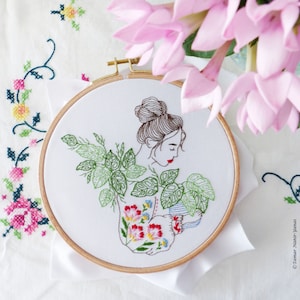May include: An embroidery hoop featuring a woman with a bun, surrounded by green leaves and flowers. The woman is holding a small pot with a plant. The embroidery is on white fabric, and the hoop is made of wood.