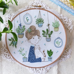 May include: Embroidered hoop art featuring a woman with brown hair and a cat, surrounded by hanging potted plants. The design is done in shades of green, blue, and brown.