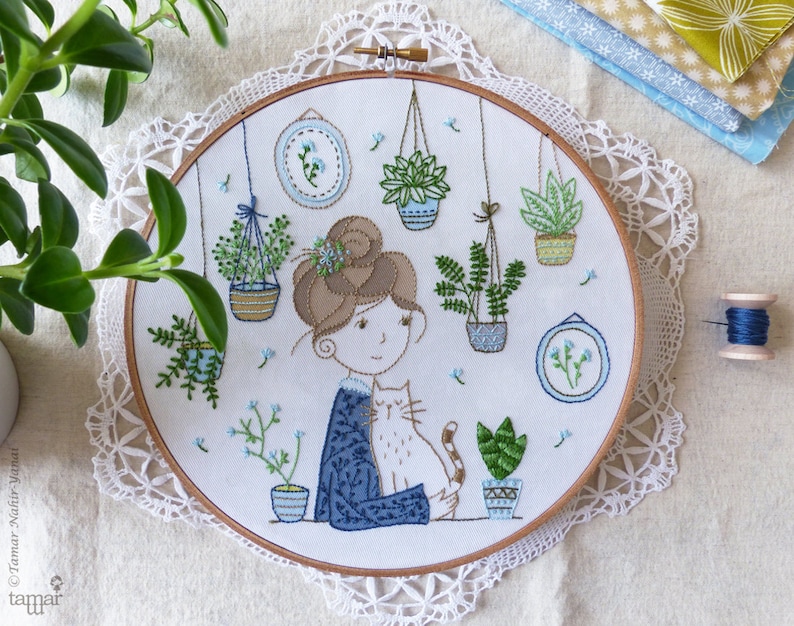 May include: Embroidered hoop art featuring a woman with brown hair and a cat, surrounded by hanging potted plants. The design is done in shades of green, blue, and brown.