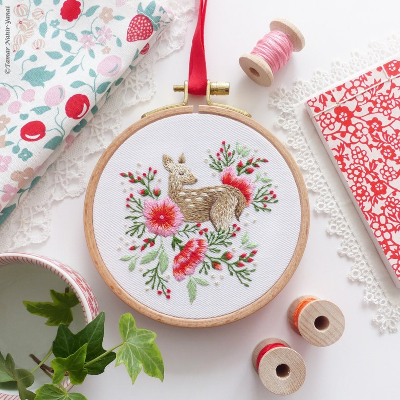 May include: Embroidered hoop art featuring a fawn surrounded by pink and red flowers. The hoop is made of wood and has a white fabric background.