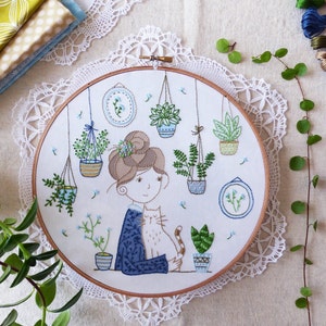 May include: Embroidered hoop art featuring a woman with brown hair and a cat, surrounded by potted plants and flowers. The hoop is white with a brown wooden frame.