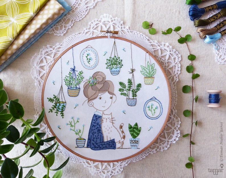 May include: Embroidered hoop art featuring a woman with brown hair and a cat, surrounded by potted plants and flowers. The hoop is white with a brown wooden frame.