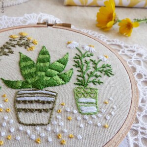 May include: Embroidered hoop art featuring green plants in pots. The plants include a succulent and a daisy-like flower. The embroidery is on a beige fabric, with a wooden hoop. Small white and yellow embroidered accents are scattered around the plants.