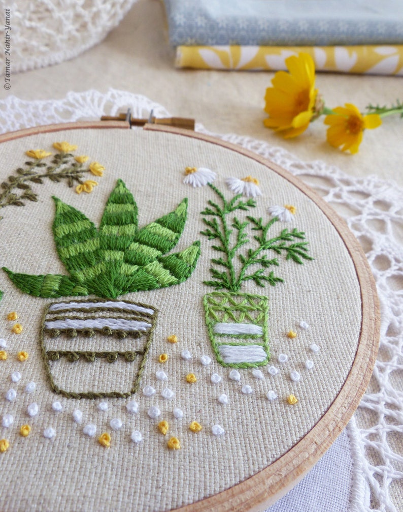 May include: Embroidered hoop art featuring green plants in pots. The plants include a succulent and a daisy-like flower. The embroidery is on a beige fabric, with a wooden hoop. Small white and yellow embroidered accents are scattered around the plants.