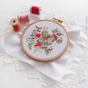 May include: Embroidered hoop art featuring a brown deer with white spots surrounded by pink and red flowers and green leaves on a white fabric background.