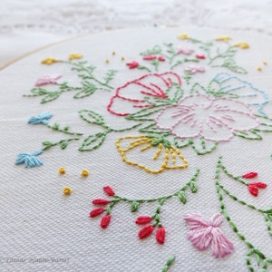 以下が含まれることがあります： 白い刺繍枠にカラフルな花のデザインが施されたクローズアップ。デザインには、ピンク、ブルー、イエロー、レッドの花と緑の葉が描かれています。