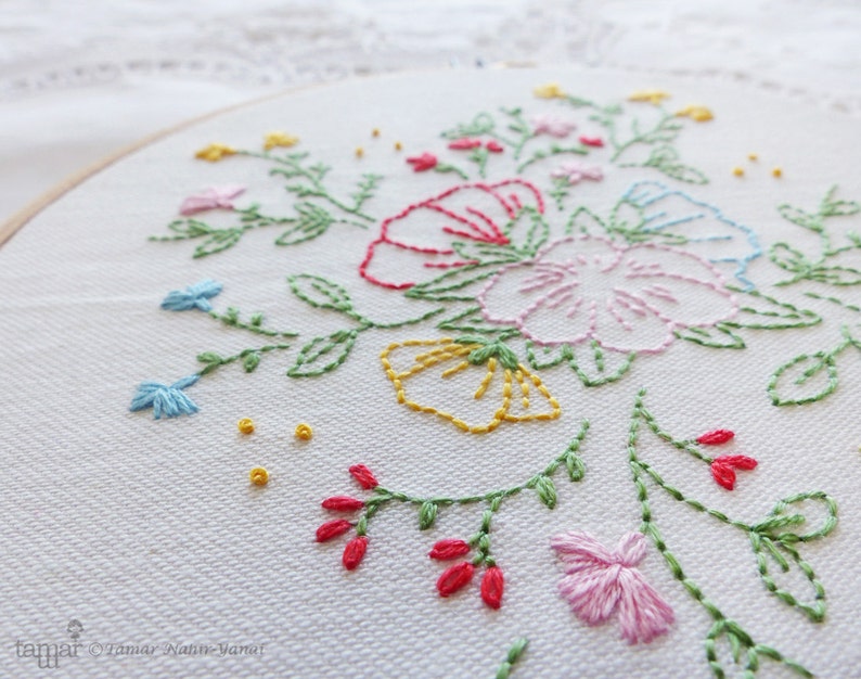 以下が含まれることがあります： 白い刺繍枠にカラフルな花のデザインが施されたクローズアップ。デザインには、ピンク、ブルー、イエロー、レッドの花と緑の葉が描かれています。