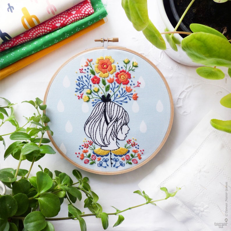 Op de afbeelding: Borduurwerk in een ronde hoepel met een meisje met een boeket kleurrijke bloemen op haar hoofd. De achtergrond is lichtblauw met witte regendruppels.