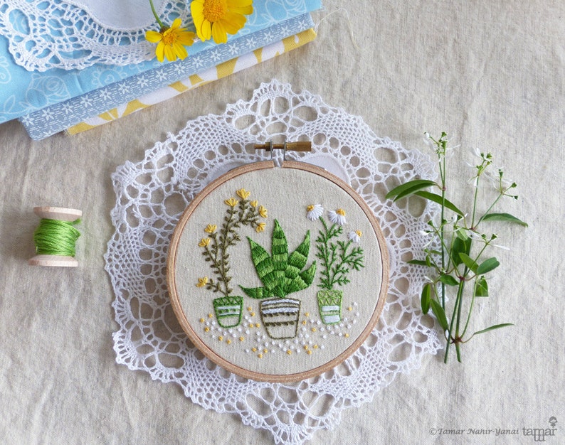 May include: A handcrafted embroidery hoop featuring three potted plants with green leaves and yellow flowers. The plants are in small pots with striped and dotted designs. The embroidery is set on a beige fabric, surrounded by a wooden hoop and a white lace doily.