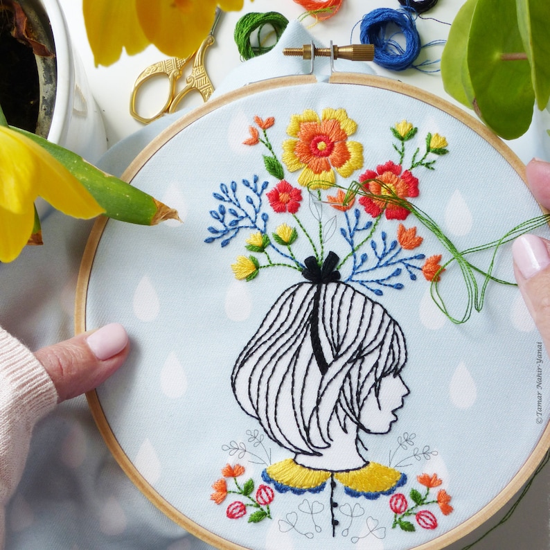 Op de afbeelding: Borduurkunst op een hoepel met een zwart-witte lijntekening van een persoon met een boeket kleurrijke bloemen op hun hoofd. De achtergrond is een lichtblauwe stof met witte stippen.